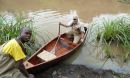 A man sits in a canoe while another holds the tip of the canoe from the shore of a small river