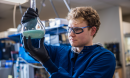 A scientist wearing protective lab gear holds up a flask containing a blue liquid.