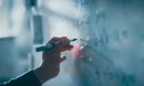 Close-up image of a hand writing equations on a white board.