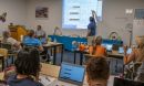 An instructor points to something projected on a screen while teachers look on from tables with laptop computers on them.
