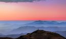 A colorful sunset and layers of blue mountaintops backdrops the Cerro Tololo Inter-American Observatory. A small profile image of the Víctor M. Blanco 4-meter Telescope is visible atop Cerro Tololo itself.