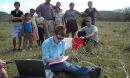 Photo of Jill Bruning entering data with small group standing behind her in a  farmer's field
