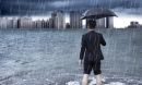 Man standing in floodwaters and holding an umbrella looks toward a city skyline.
