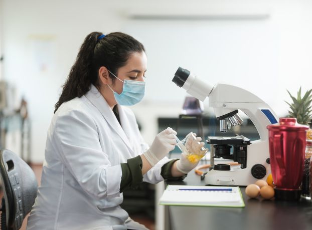 A latin female lab student wearing gloves and mask, sitting and holding a flask with an egg inside.