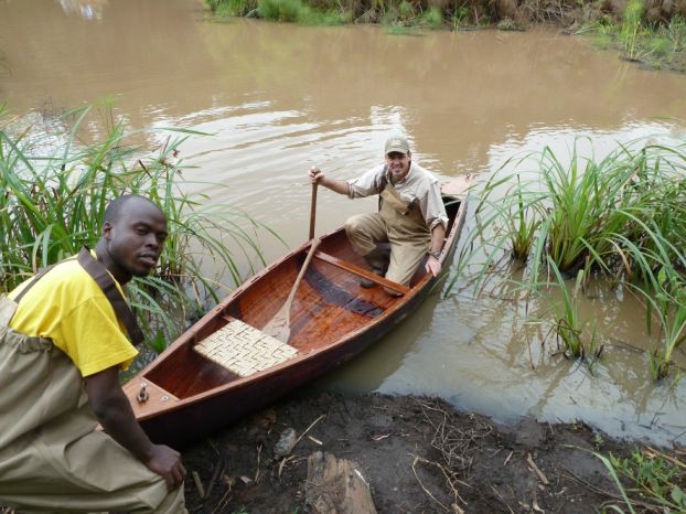 A man sits in a canoe while another holds the tip of the canoe from the shore of a small river