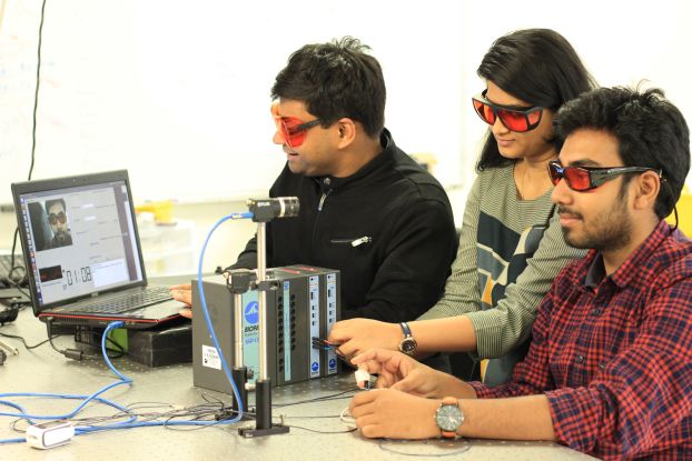 Three students wearing orange glasses, seated in front of a computer and a monitoring device.