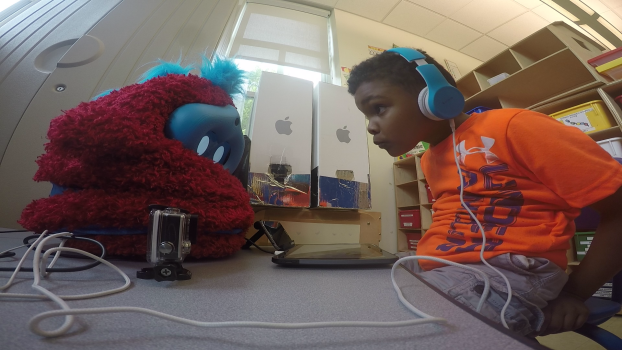 A child wearing headphones looks attentively at a furry red robot.