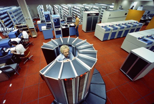 A man stands among supercomputer racks in a 1970's era room with orange-red tile floors.
