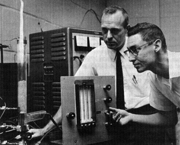 A black-and-white photo of two researchers interacting with a piece of equipment in a research lab.