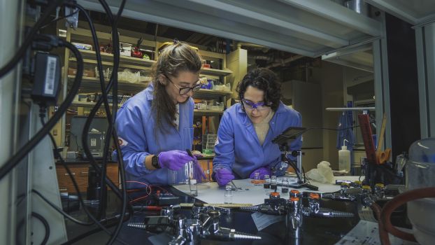 Two women in blue lab coats and purple gloves bend over a lab bench.