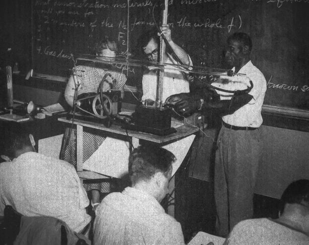 A black-and-white photo of teachers standing at the front of a classrom with an experimental set-up.