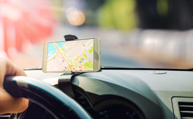 Smart phone attached to the dashboard of a car displaying a GPS navigation map with a hand on the steering wheel.