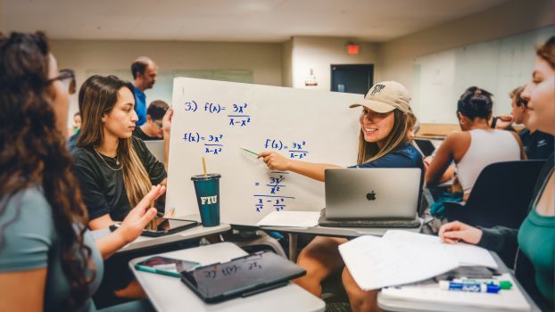 Students sitting at desks pulled together closely look at math equations on a white board.