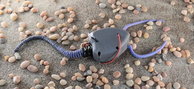 A small robot with a purple mechanical tail sits on sand and rocks.