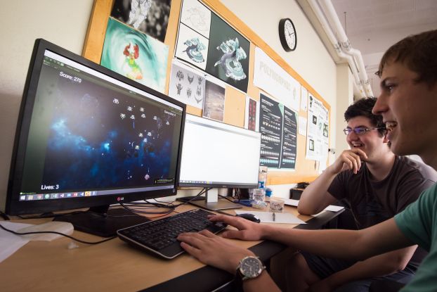 Smiling students sit at a computer, playing a video game they designed.