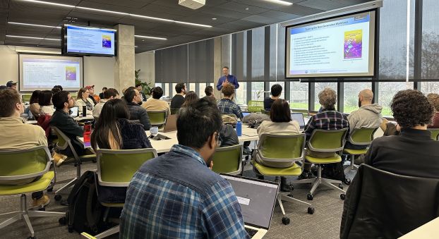 A man speaking in front of a room of seated graduate students. He is standing next to a PowerPoint with a slide titled "Training the Next Generation."