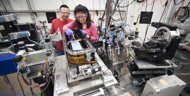 Two researchers in protective eyewear adjust an apparatus in a lab full of metal equipment and wiring.