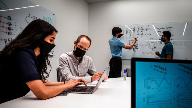 Student with scientist sitting in front of laptop together while two students in the background diagram systems at the white board