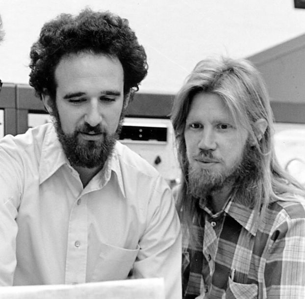 A black-and-white 1970s-era photo of two men looking at data printed on a ream of paper.