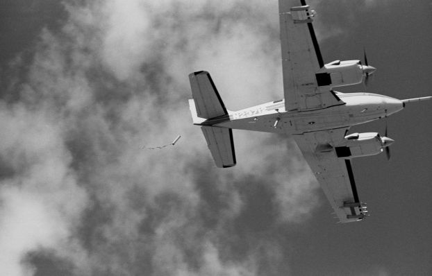 A black-and-white photograph of the underside of a plane in flight that has just dropped a small tube.