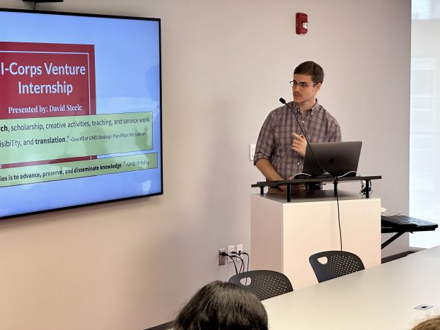 A person stands at a lectern giving a presentation. The screen behind them says "I-Corps Venture Internship."