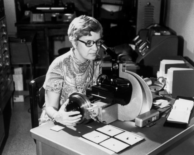 A black-and-white 1970s-era photo of a woman peering into the viewfinder of a large piece of laboratory equipment.