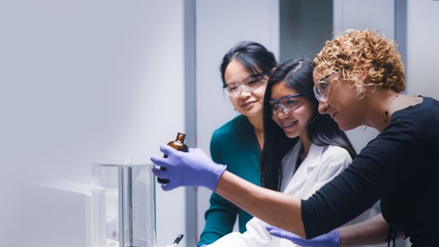 Interns in lab coats and protective eyewear looking at bottled sample held by scientist.