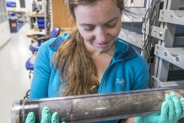 A researcher holds a large glass tube containing basalt in her gloved hands.