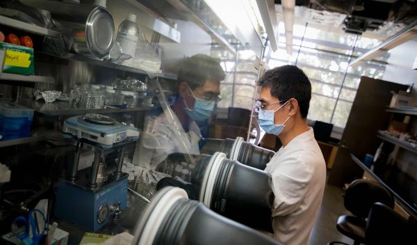 A man in a lab working on something behind a protective glass shield.