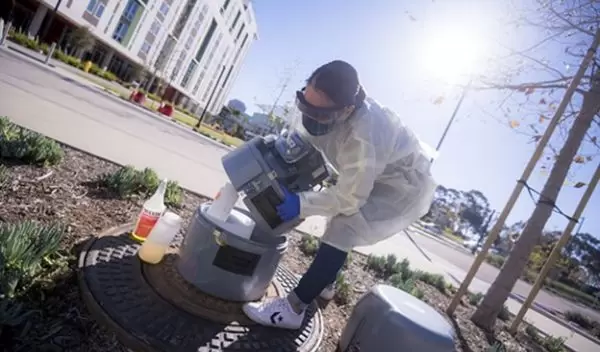 A researcher who is wearing protective labwear handles a wastewater sampler outdoors.