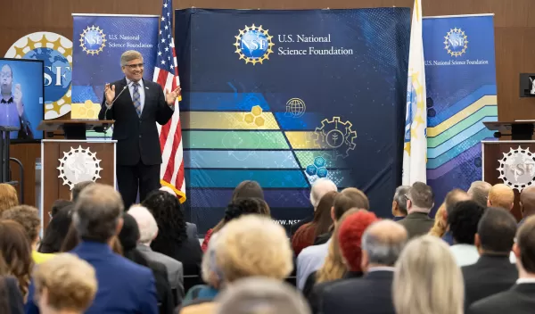 man on a stage speaking in front of an audience clothed in a suit