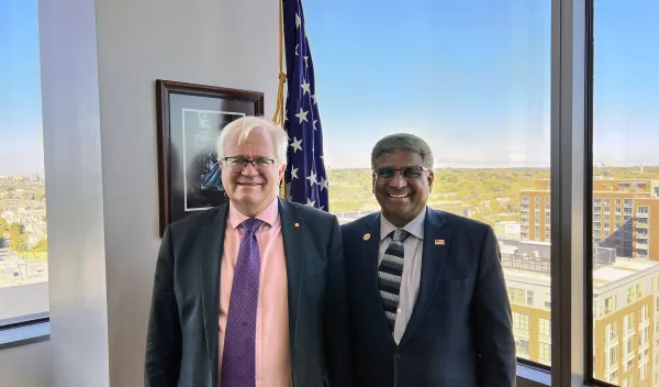 two men standing in front of a U.S. flag, both wearing suit and ties