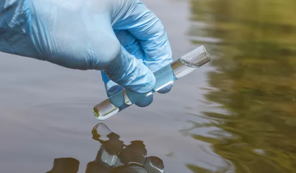 Hand wearing a blue latex glove holding a test tube over a body of water.