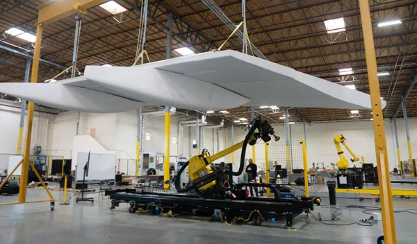 A white aircraft wing suspended above a mobile robot with a large yellow arm in an open hangar space.