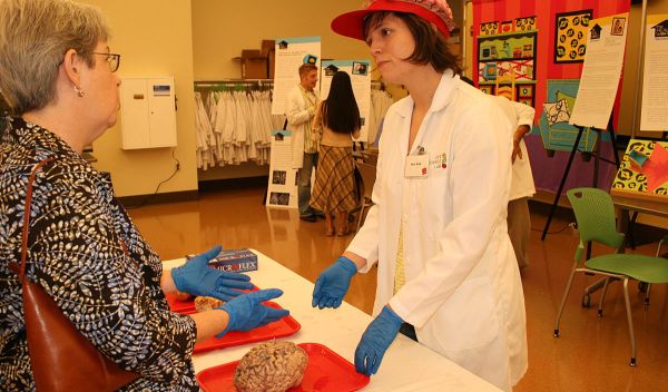 Photo of Alexis Webb, in brain hat and in front of an actual brain, answering a visitor's question.