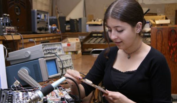 a young woman holding a circuit board and next to table with electronics.