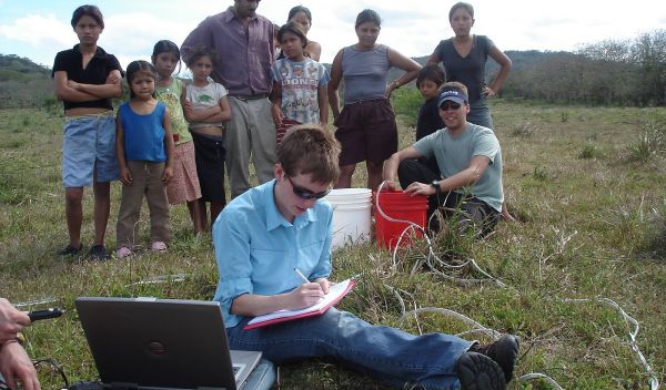 Photo of Jill Bruning entering data with small group standing behind her in a farmer's field