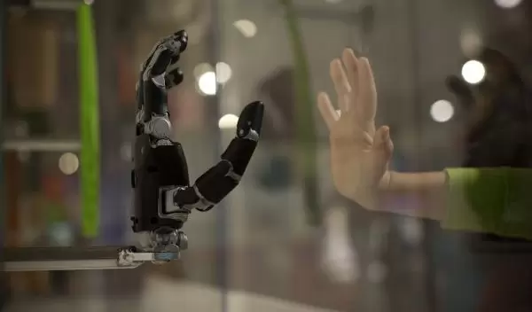 Human hand held up to a plate of glass, with a robotic hand mirroring the same gesture on the other side of the glass
