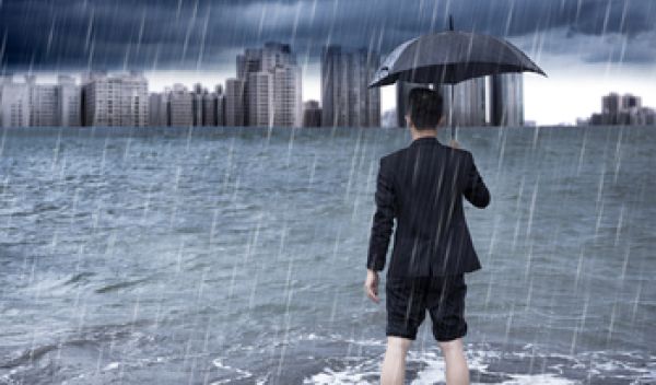 Man standing in floodwaters and holding an umbrella looks toward a city skyline.