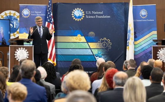 man on a stage speaking in front of an audience clothed in a suit