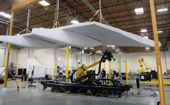 A white aircraft wing suspended above a mobile robot with a large yellow arm in an open hangar space.