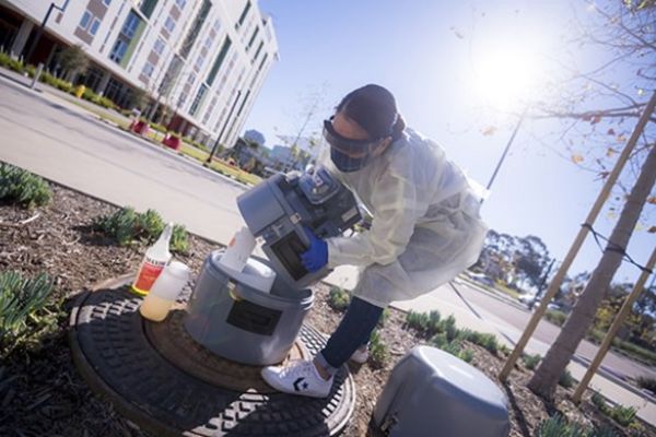 A researcher who is wearing protective labwear handles a wastewater sampler outdoors.
