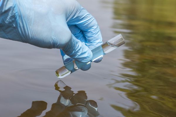 Hand wearing a blue latex glove holding a test tube over a body of water.