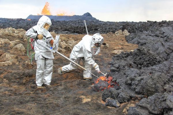 two men standing in a volcanic field wearing protective suits and masks