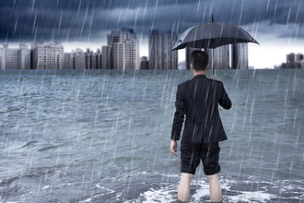 Man standing in floodwaters and holding an umbrella looks toward a city skyline.